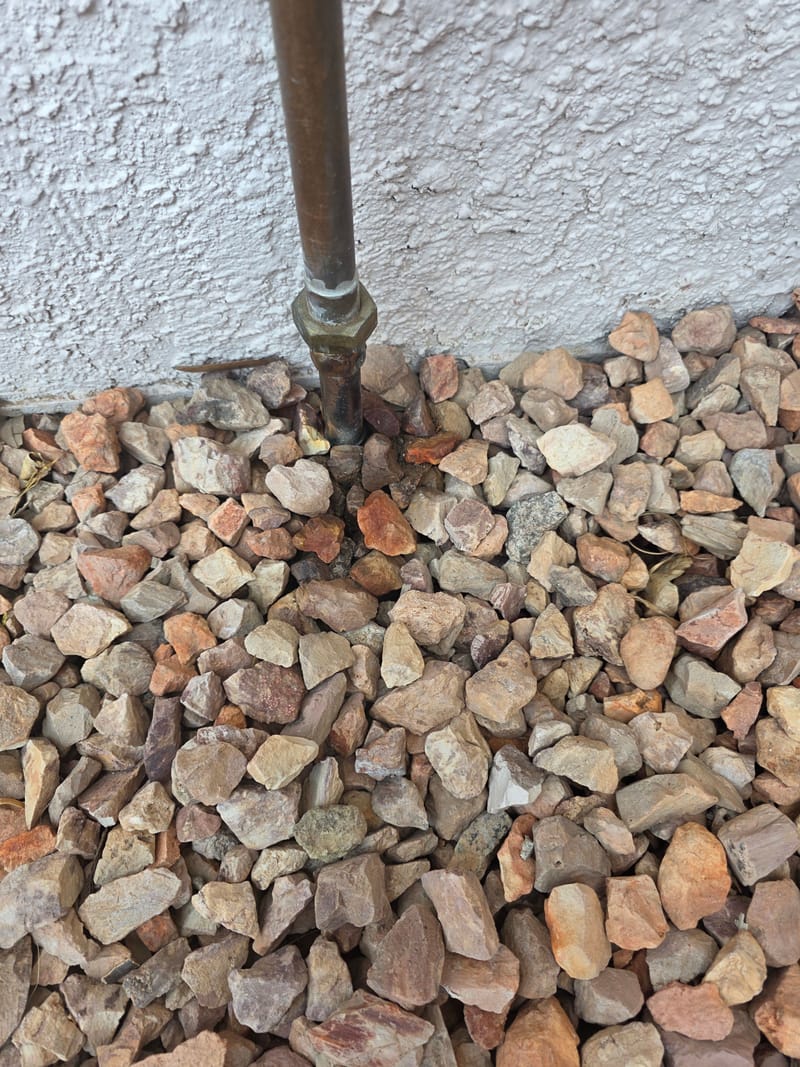 Copper water line and shutoff rising from gravel beside a stucco wall